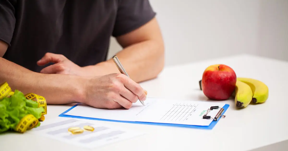 A person writes on a clipboard at a table with bananas, an apple, lettuce, a measuring tape, and pills nearby—reflecting the holistic approach to wellness offered by Choices Psychotherapy and mental health services.