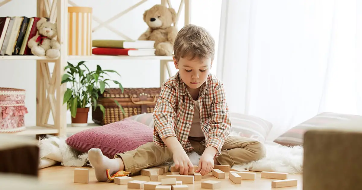 A young child sits cross-legged on the floor playing with wooden blocks, surrounded by cushions, shelves with books, plants, and stuffed animals in a bright room designed to support mental health services.