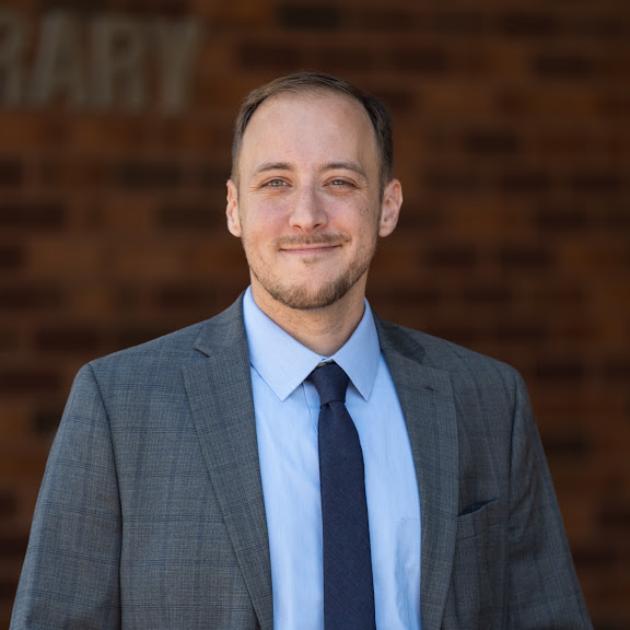 A man in a gray suit, blue shirt, and navy tie stands outside in front of a brick wall, smiling slightly at the camera—introducing Keith Block, LICSW.