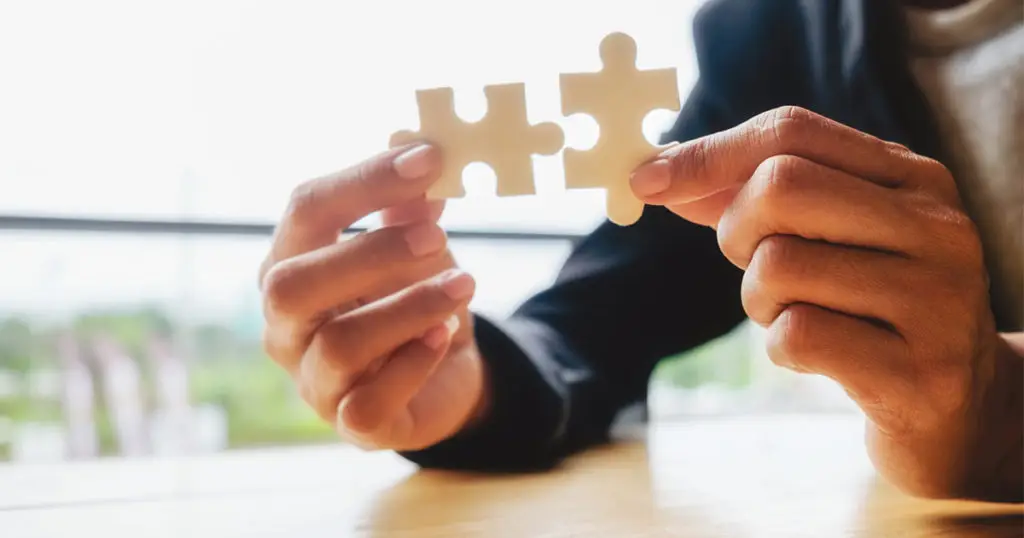A person in business attire holds two puzzle pieces close together on a wooden table, symbolizing how Choices Psychotherapy connects individuals to effective mental health services, with a blurred background emphasizing focus.