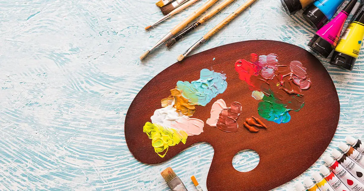 A wooden artist palette with various paint colors, tubes of paint, and paintbrushes arranged on a textured light blue surface, reflecting creative expression supported by Choices Psychotherapy’s mental health services.
