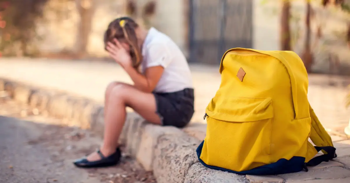 A young girl sits on a curb with her head in her hands next to a large yellow backpack.