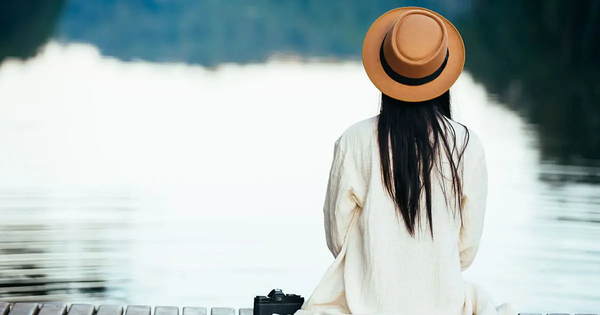A person wearing a brown hat and light-colored clothing sits on a dock facing a calm body of water, reflecting in peaceful solitude—much like the self-discovery supported by Choices Psychotherapy and mental health services. A camera rests beside them.