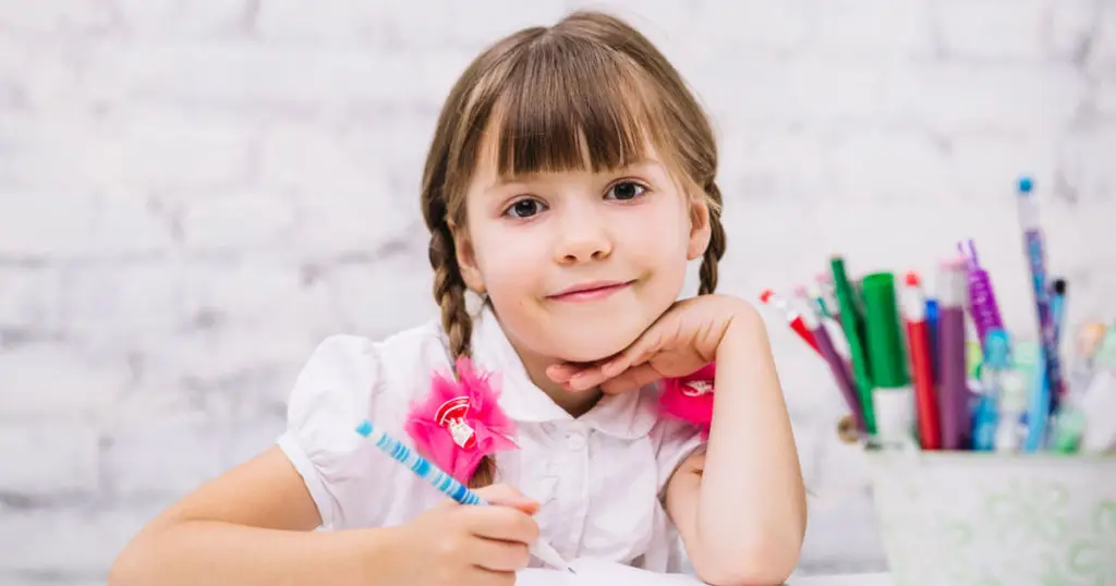 Young girl with braided hair sits at a desk, holding a pencil and smiling, with a container of colorful pens and pencils in the background—reflecting the positive impact of Choices Psychotherapy and mental health services.