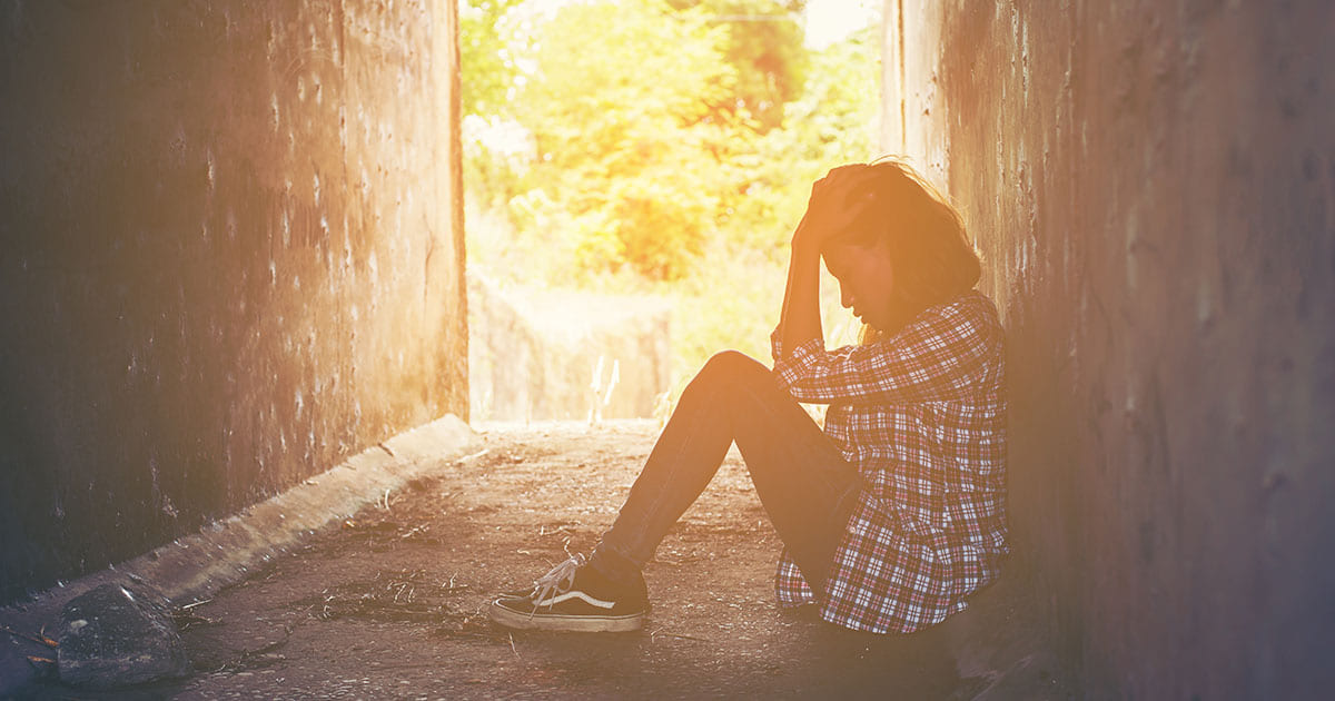 A person sits on the ground in a narrow, dimly lit passageway, holding their head with both hands, appearing distressed. Sunlight shines in from the opening behind them—reminding us of the help Choices Psychotherapy and mental health services provide.