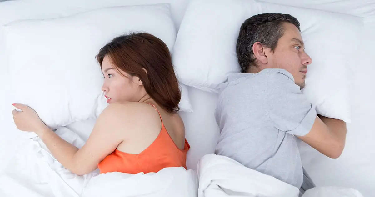A woman and a man lie in bed back-to-back, looking away from each other with neutral expressions, highlighting the silent strain that underscores the need for mental health services like those at Choices Psychotherapy.