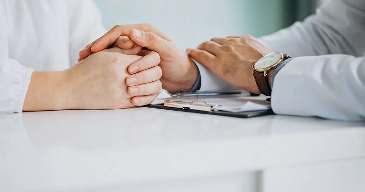 A doctor sits across from a patient, holding their hands in comfort, with a clipboard and pen on the white desk between them, reflecting the compassionate approach of Choices Psychotherapy’s mental health services.