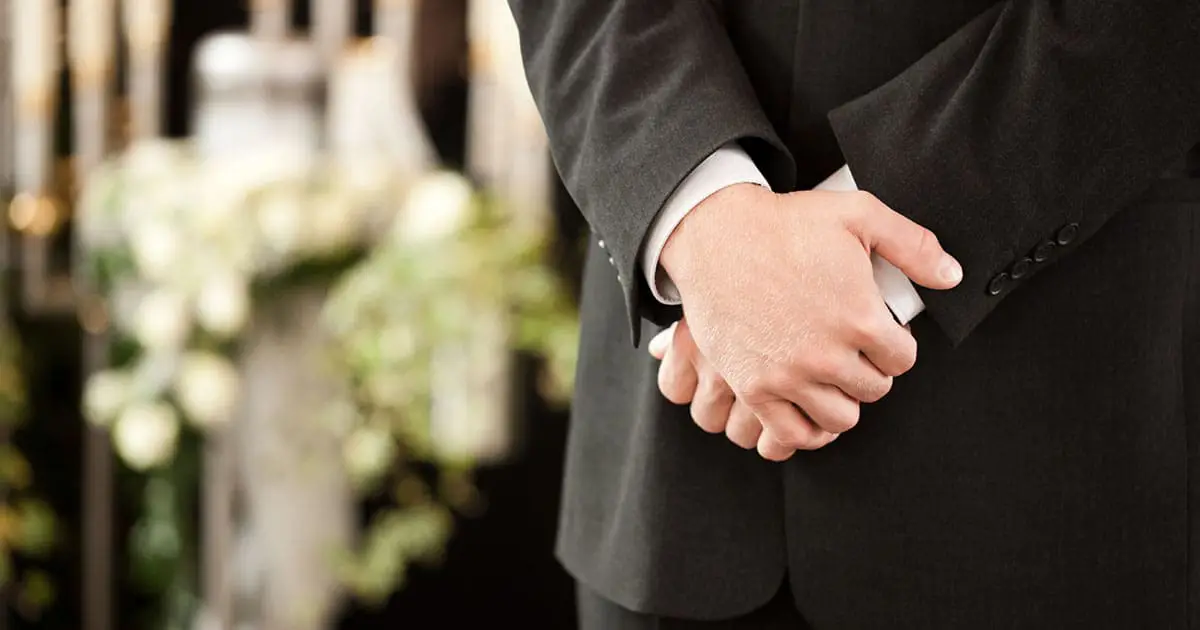 A person in a black suit stands with hands clasped at a formal event, with blurred floral arrangements and candles in the background—reflecting on support found through Choices Psychotherapy and mental health services.