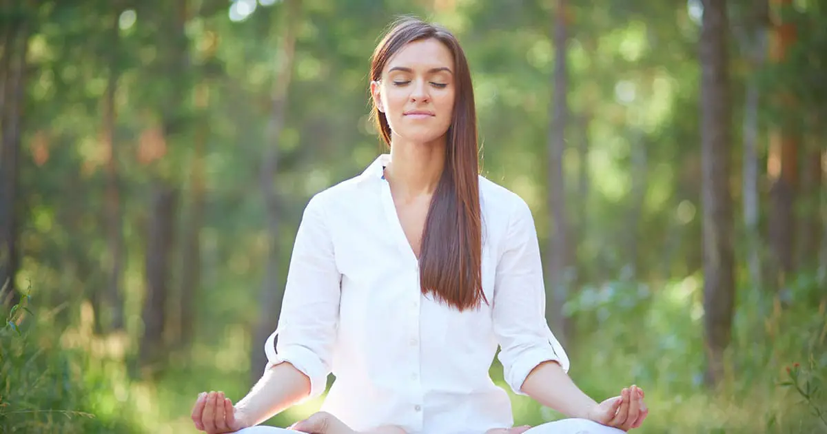 A woman in a white shirt sits cross-legged on the grass in a forest, meditating with her eyes closed, embracing tranquility—a moment inspired by Choices Psychotherapy and support for mental health services.