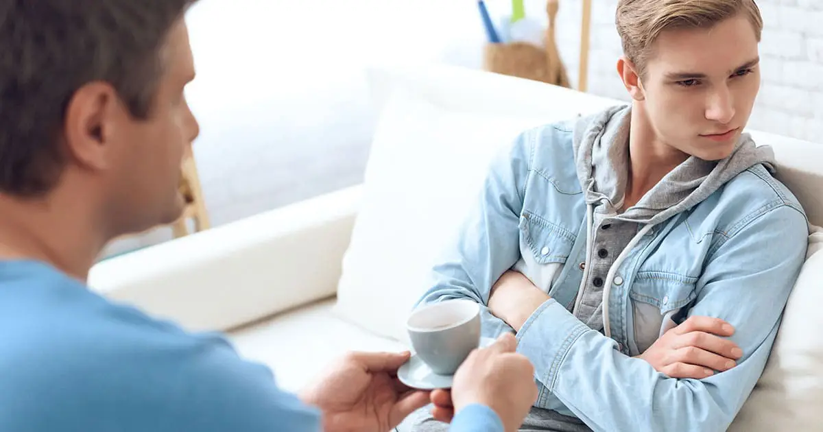 An adult offers a cup to a teenage boy sitting on a couch with his arms crossed, looking away with an unhappy expression—illustrating the importance of mental health services like Choices Psychotherapy in supporting teens.