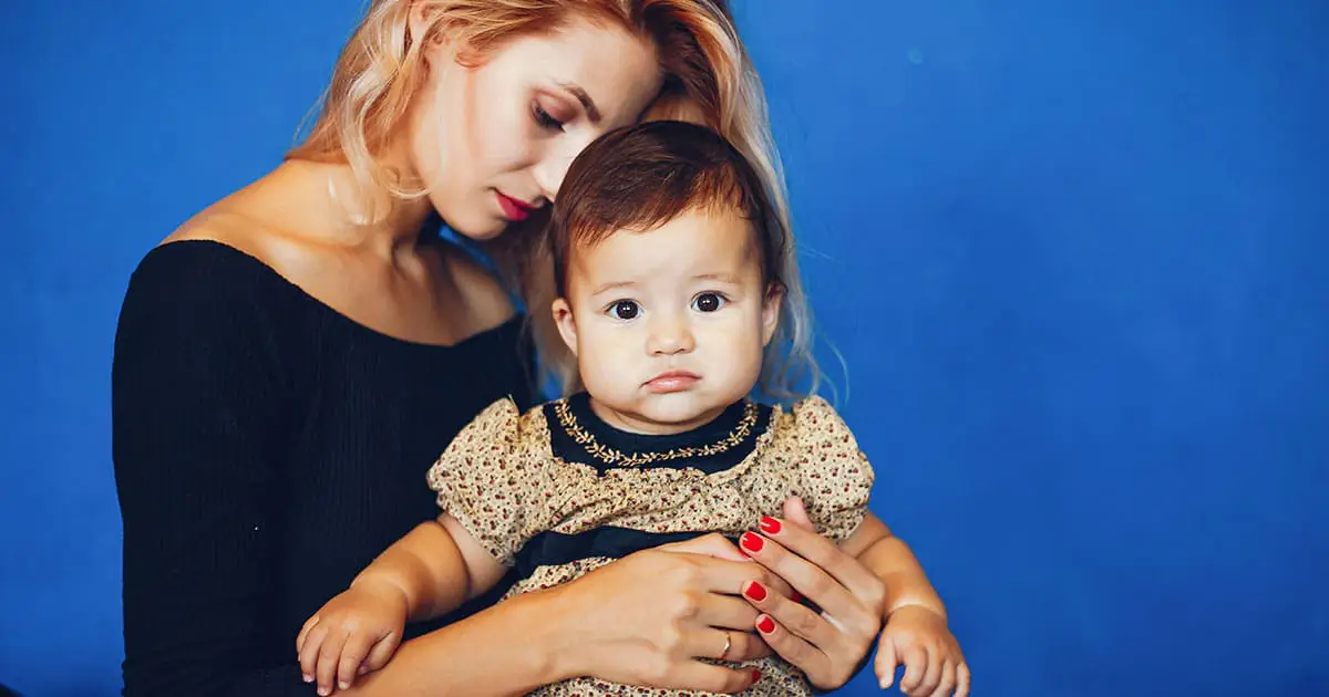 A woman with blonde hair holds a toddler wearing a patterned dress, both facing the camera, against a solid blue background, highlighting the supportive environment fostered by Choices Psychotherapy.