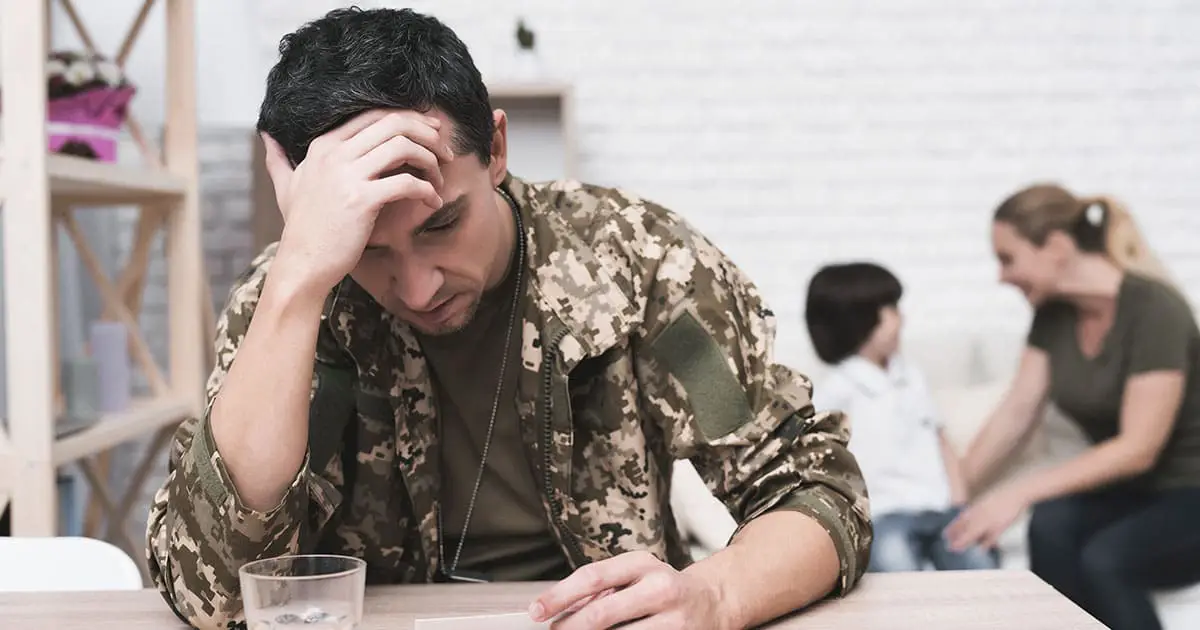 A man in military uniform sits at a table with his head in his hand, while a woman and child talk in the background, highlighting the importance of mental health services like Choices Psychotherapy for families.