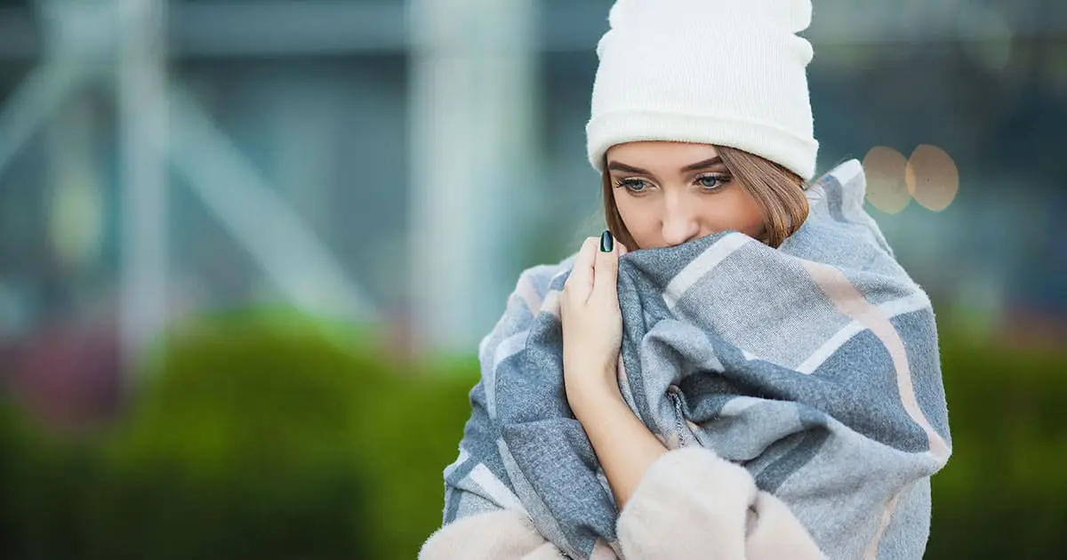 A woman wearing a white knit hat and wrapped in a gray plaid blanket stands outdoors, looking down with her hands holding the blanket close to her face—reflecting the warmth and care found at Choices Psychotherapy.