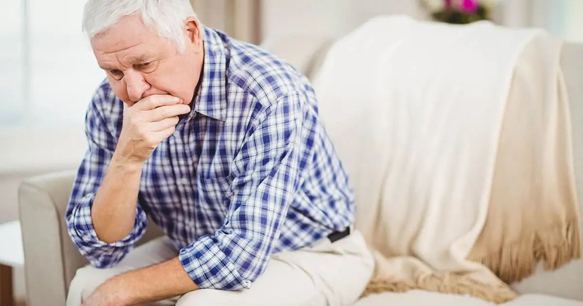 An older man in a plaid shirt sits on a couch, leaning forward with his hand over his mouth, appearing deep in thought or concerned—perhaps reflecting on seeking mental health services like those offered at Choices Psychotherapy.