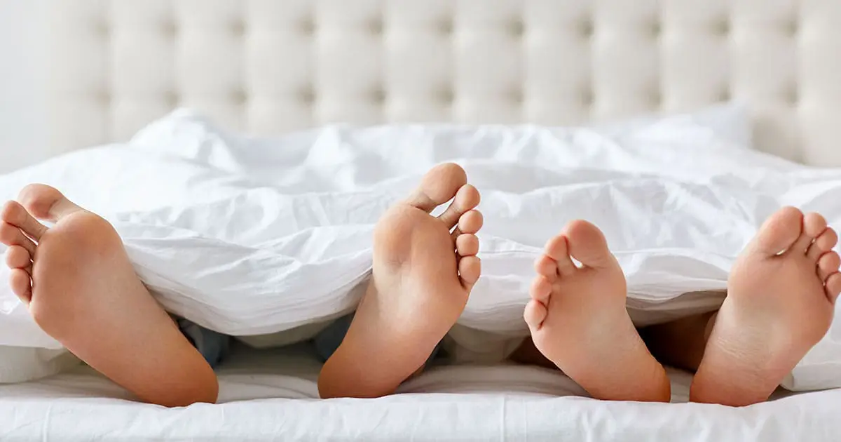 Two pairs of bare feet stick out from under a white blanket on a bed with a tufted headboard, reflecting the comfort and trust that Choices Psychotherapy and mental health services strive to provide.