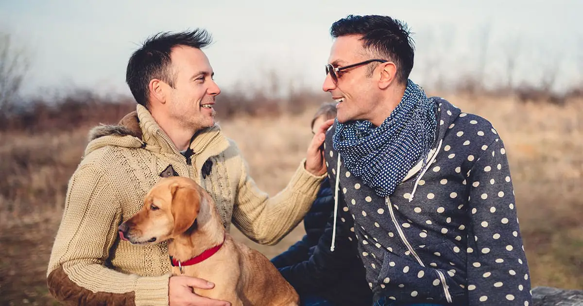 Two men sit outdoors on dry grass, one holding a dog. They face each other, smiling and talking in winter clothing, sharing a moment that reflects the supportive connection found through Choices Psychotherapy and mental health services.
