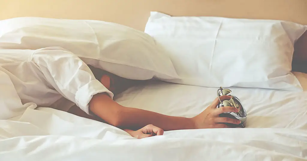 Person lying in bed under white covers, reaching out one arm to turn off an alarm clock—starting the day with self-care and support from Choices Psychotherapy, where mental health services are tailored to each individual's needs.
