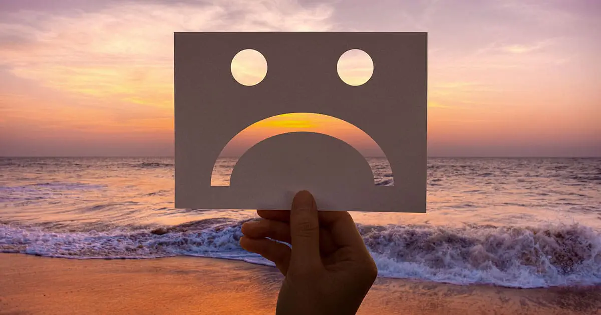 A hand holds a paper cutout of a sad face in front of an ocean at sunset, symbolizing the importance of seeking mental health services like Choices Psychotherapy for support during difficult times.