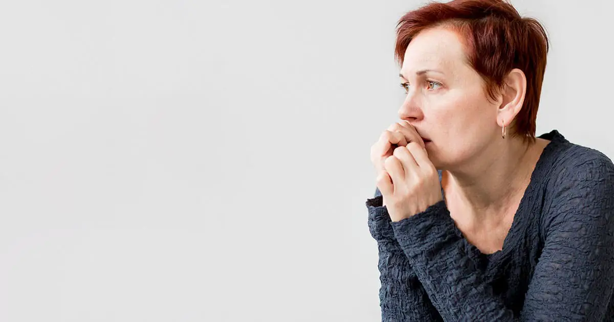 A middle-aged woman with short hair wearing a textured dark sweater is sitting and looking to the side, appearing worried or anxious, with her hands near her mouth—illustrating the importance of mental health services like Choices Psychotherapy.